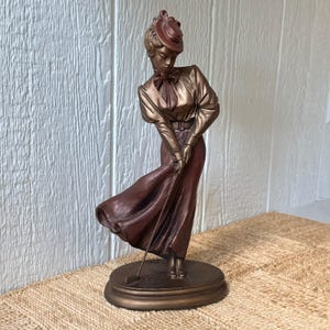 May include: A bronze-toned figurine of a person wearing a long-sleeved blouse, a long skirt, and a hat, standing on an oval base. The figure is holding a golf club with both hands in a downward position, with the skirt appearing to flow to one side.