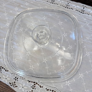 May include: A clear, square glass lid with a central handle. The lid has rounded corners and a raised rim. The handle is a small, circular knob. The lid is designed for cookware.