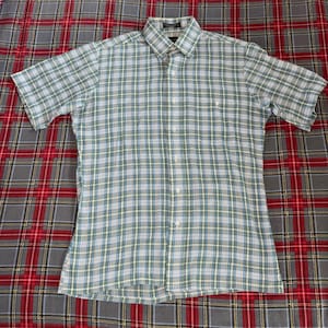 Puede incluir: Camisa de manga corta con estampado de cuadros verdes, azules y blancos. La camisa tiene cuello abotonado y un bolsillo en el pecho. La tela es ligera. La camisa está sobre una superficie a cuadros.