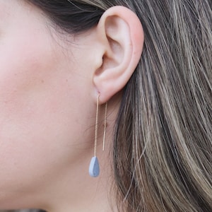 Puede incluir: Un par de delicados pendientes de cadena dorada con un colgante en forma de lágrima azul claro. Los pendientes se muestran puestos en la oreja de una persona.
