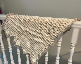 May include: A square crochet blanket featuring a textured cream-colored center with a repeating bobble stitch pattern. The blanket is finished with a decorative scalloped border in a contrasting light beige yarn, creating a distinct edge.