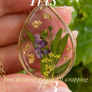 May include: A gold-colored pendant necklace with a teardrop-shaped resin charm. The charm contains dried purple and yellow flowers, green leaves, and gold flakes. The word "Iris" is at the top, and "February birth flower" is at the bottom.
