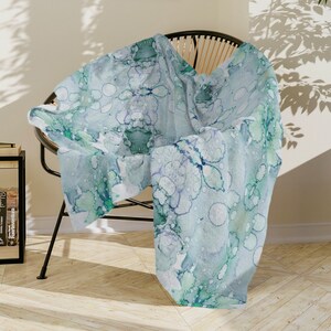 May include: A soft, light blue throw blanket with a white and green abstract pattern. The blanket is draped over a wicker chair in a room with a wooden floor.