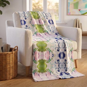 May include: A soft throw blanket featuring a repeating abstract watercolor pattern in shades of lime green, soft pink, and deep navy blue on a white background, draped over a white upholstered armchair with wooden legs in a bright room.