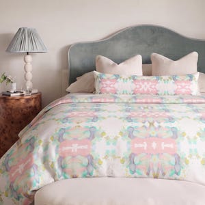 May include: A bed featuring a duvet cover and a long bolster pillow with a repeating abstract pattern in pastel pink, light blue, and soft yellow. The bed has a light blue velvet headboard, two cream pillows, and a cream-colored pleated bench.