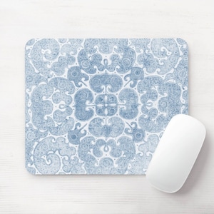 May include: A rectangular mousepad with a blue and white paisley design. The pattern is symmetrical and features swirling floral and geometric shapes. A white computer mouse is positioned on the right side of the mousepad.