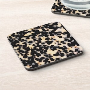 May include: A set of two square coasters with a black and white mottled pattern reminiscent of tortoiseshell. The coasters have a black border.