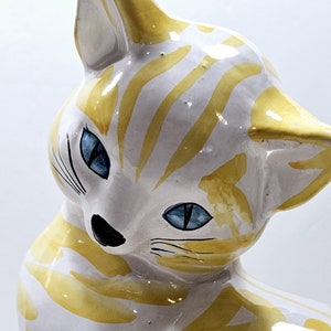 Pode incluir: Uma figura de cerâmica de um gato com listras amarelas e brancas. O gato tem olhos azuis e um nariz preto.