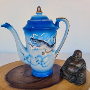 May include: A blue and white porcelain teapot with a dragon design. The teapot has a gold handle and spout. It is sitting on a wooden surface next to a small bronze statue of a Buddha.