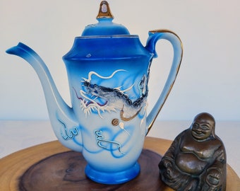 Könnte beinhalten: Eine blaue und weiße Porzellan-Teekanne mit einem Drachen-Design. Die Teekanne hat einen goldenen Henkel und Ausguss. Sie steht auf einer Holzoberfläche neben einer kleinen Bronzestatue eines Buddha.