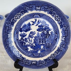 May include: A blue and white porcelain plate with a willow pattern design. The plate features a stylized landscape with a bridge, a pagoda, and a willow tree. The plate is on a black stand.