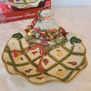 May include: A ceramic serving dish shaped like Santa Claus's bag, with a Santa Claus figurine peeking out of the top. The bag is decorated with a green, red, and gold pattern of fruits, leaves, and vines. The dish is labeled "Renaissance Santa".