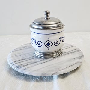 May include: A white and blue ceramic jar with a metal lid and base, decorated with blue scroll and diamond patterns. The jar sits on a round marble tray with gray veining. The jar has a small metal knob on top.