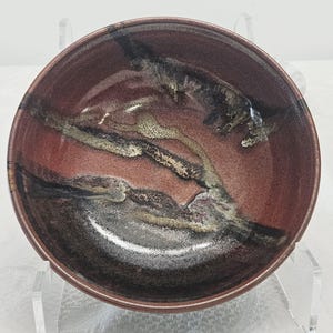 Puede incluir: Un cuenco de cerámica hecho a mano con exterior e interior marrón rojizo. El interior presenta un diseño único con vetas negras, grises y beige. El cuenco es redondo y tiene un acabado liso y brillante.