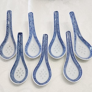 Könnte beinhalten: Ein Set aus sieben weißen Keramik-Suppenlöffeln mit einem blauen geometrischen Muster entlang der Griffe und einem blauen Blumenmuster in der Mitte der Löffelschale. Die Löffel haben einen geschwungenen Griff und eine tiefe, runde Laffe.