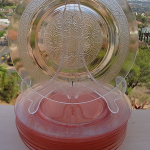 May include: A stack of vintage pink depression glass plates with a floral design. The top plate is clear glass with a raised floral pattern. The plates are stacked on a white surface.