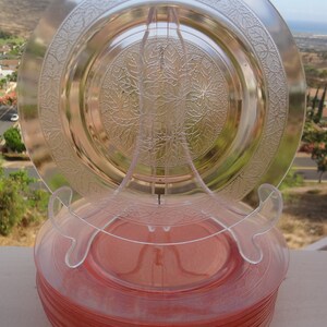 May include: A stack of 10 pink depression glass plates with a clear glass plate on top. The plates have a floral design.