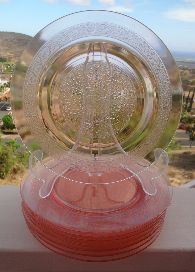 May include: A stack of 10 pink depression glass plates with a clear glass plate on top. The plates have a floral design.