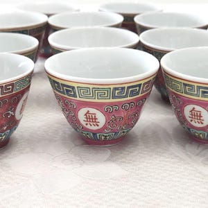 May include: A set of small tea cups with a white interior and a red exterior. The cups are decorated with gold, green, and blue patterns, and feature a Chinese character in a white circle. The cups are displayed on a white surface.