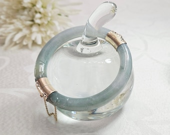 May include: A light green jadeite bangle bracelet with gold-colored clasps and a delicate chain. The bracelet is displayed on a clear glass object, with a white flower in the background. The jadeite has a translucent quality.