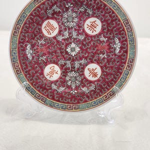 May include: A decorative, round ceramic plate with a red background and intricate white floral and scrollwork design. The plate features four Chinese characters and a geometric border. It is displayed on a clear stand.