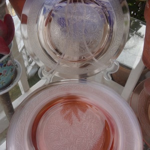 May include: Two vintage glass plates with a floral design. The top plate is clear glass with a frosted floral pattern. The bottom plate is pink glass with a frosted floral pattern.