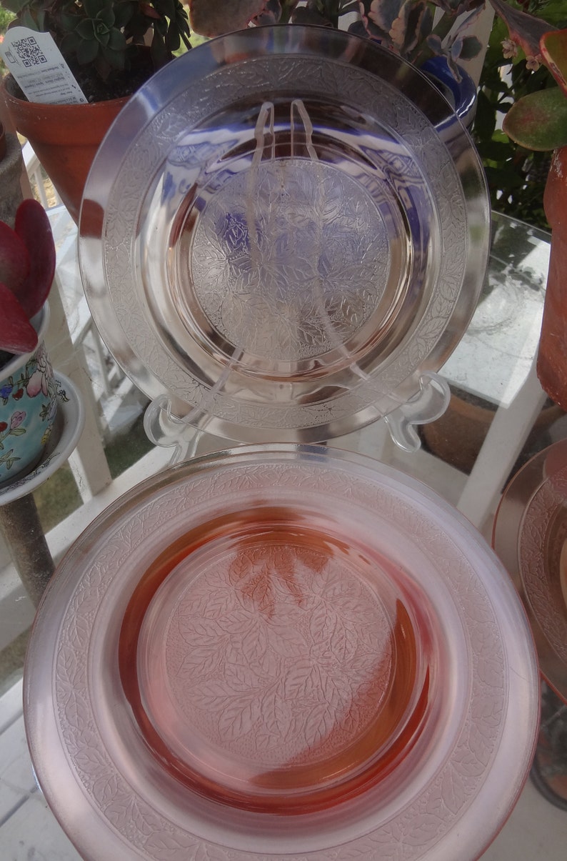May include: Two vintage glass plates with a floral design. The top plate is clear glass with a frosted floral pattern. The bottom plate is pink glass with a frosted floral pattern.