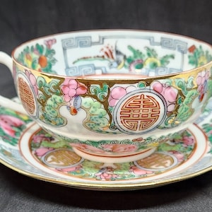 May include: A white porcelain teacup and saucer set with a colorful floral and geometric design. The cup and saucer feature a gold trim and a red Chinese character.