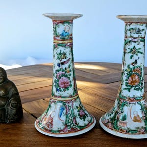 Könnte beinhalten: Zwei weiße Porzellan-Kerzenhalter mit floralem und figürlichem Design. Die Kerzenhalter stehen auf einem Holztisch mit einer Bronzestatue von Buddha.