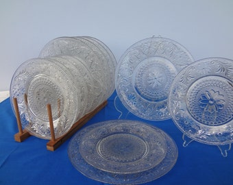May include: A set of clear glass dinner plates with a decorative floral pattern. The plates are stacked on a wooden rack and displayed on a blue surface.