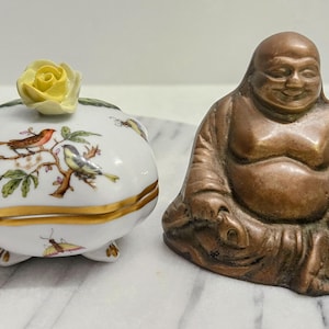 May include: A white porcelain trinket box with a yellow rose on top and a gold trim, decorated with birds and insects. Next to it is a bronze-colored Buddha statue.