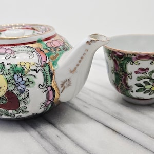 May include: A white porcelain teapot and teacup set with a colorful floral design. The teapot has a lid and spout, and the teacup features a bird and floral pattern. Gold trim accents the edges.