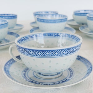 May include: A set of white porcelain teacups and saucers with blue decorative trim. The teacups have a delicate, pierced design and a handle. The saucers and teacups have a matching blue geometric border. The set is arranged on a white surface.