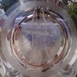 May include: A clear glass plate with a delicate floral pattern etched into the surface. The plate has a raised rim and a slightly scalloped edge.