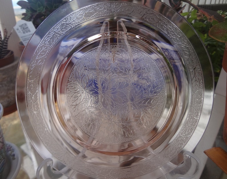 May include: A clear glass plate with a delicate floral pattern etched into the surface. The plate has a raised rim and a slightly scalloped edge.