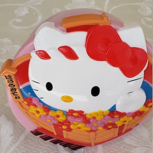 Può includere: Un salvadanaio rosa Hello Kitty con un fiocco rosso, un viso bianco e un design floreale. Il salvadanaio ha una striscia blu e arancione e il testo "Hello Kitty" sul lato.