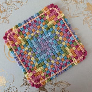 May include: A square, handwoven coaster with a colorful, checkered pattern. The coaster features a variety of pastel colors, including pink, blue, yellow, and green. A pink border and scalloped edge frame the design, creating a decorative and functional item.