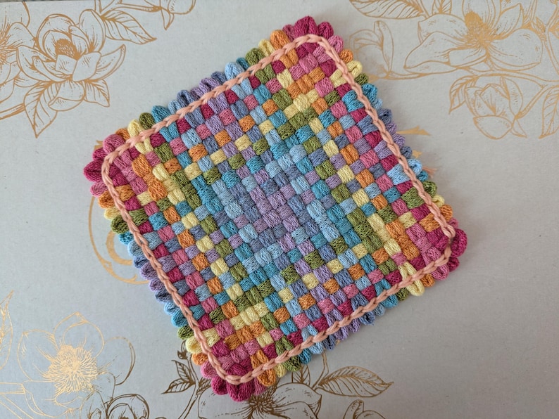 May include: A square, handwoven coaster with a colorful, checkered pattern. The coaster features a variety of pastel colors, including pink, blue, yellow, and green. A pink border and scalloped edge frame the design, creating a decorative and functional item.