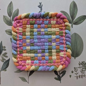 May include: A square, woven trivet in pastel colors. The trivet features a woven pattern in shades of pink, orange, yellow, green, blue, and purple. The edges are finished with a braided border in coordinating colors. The trivet is approximately 7 inches square.