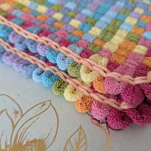 May include: Close-up of a colorful, hand-woven textile with a textured, bubble-like pattern. The piece features a variety of pastel colors, including pink, blue, yellow, and green, with a peach-colored border. The craftwork is likely a decorative item.