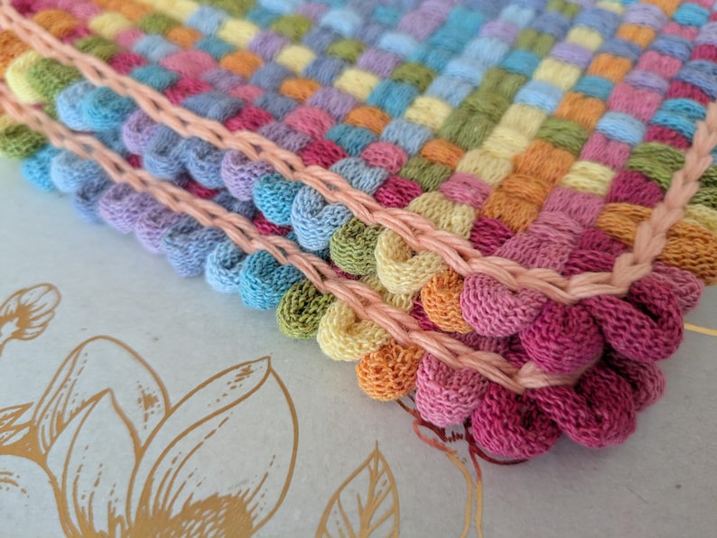 May include: Close-up of a colorful, hand-woven textile with a textured, bubble-like pattern. The piece features a variety of pastel colors, including pink, blue, yellow, and green, with a peach-colored border. The craftwork is likely a decorative item.
