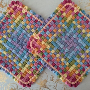 May include: Two colorful, square woven potholders. Each potholder features a vibrant mix of colors including pink, yellow, blue, and orange, with a scalloped pink border. The woven design creates a textured appearance, perfect for kitchen use.