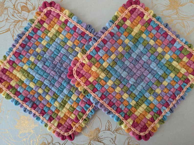 May include: Two colorful, square woven potholders. Each potholder features a vibrant mix of colors including pink, yellow, blue, and orange, with a scalloped pink border. The woven design creates a textured appearance, perfect for kitchen use.