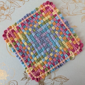 May include: A square, woven coaster in a vibrant array of colors including pink, orange, yellow, green, blue, and purple. The edges are finished with a scalloped pink trim, adding a decorative touch to this handmade item.