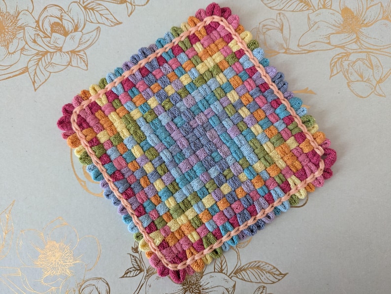 May include: A square, woven coaster in a vibrant array of colors including pink, orange, yellow, green, blue, and purple. The edges are finished with a scalloped pink trim, adding a decorative touch to this handmade item.