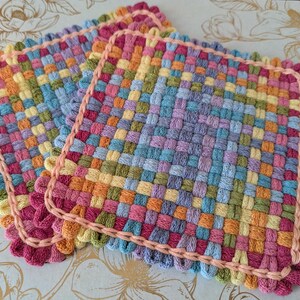 May include: A stack of colorful, woven square potholders. The potholders are made of woven yarn in a variety of colors, including pink, orange, yellow, green, blue, and purple. Each potholder has a pink border and measures approximately 8 inches square.