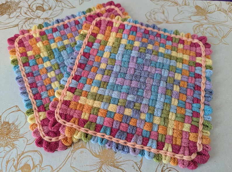 May include: A stack of colorful, woven square potholders. The potholders are made of woven yarn in a variety of colors, including pink, orange, yellow, green, blue, and purple. Each potholder has a pink border and measures approximately 8 inches square.