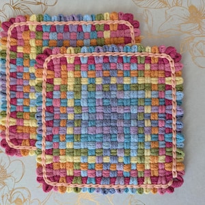 May include: Two colorful, woven square potholders. The potholders feature a woven pattern in shades of pink, orange, yellow, green, blue, and purple. Each potholder has a pink braided border. The potholders are approximately 7 inches square.
