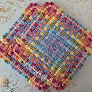 May include: Three colorful woven potholders. Each potholder features a square shape with a woven pattern in shades of pink, orange, yellow, green, and blue. The edges are finished with a scalloped pink trim.