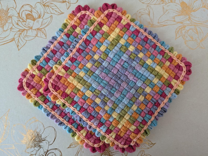 May include: Three colorful woven potholders. Each potholder features a square shape with a woven pattern in shades of pink, orange, yellow, green, and blue. The edges are finished with a scalloped pink trim.
